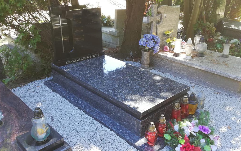 Granitowy nagrobek z wygrawerowanym krzyżem i napisem 'Spoczywajcie w pokoju', otoczony białym żwirem i lampionami, na tle innych nagrobków i drzew.