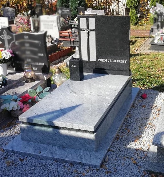 Granitowy pomnik z krzyżem i napisem 'Pokój Jego Duszy', otoczony innymi nagrobkami na cmentarzu w słoneczny dzień.