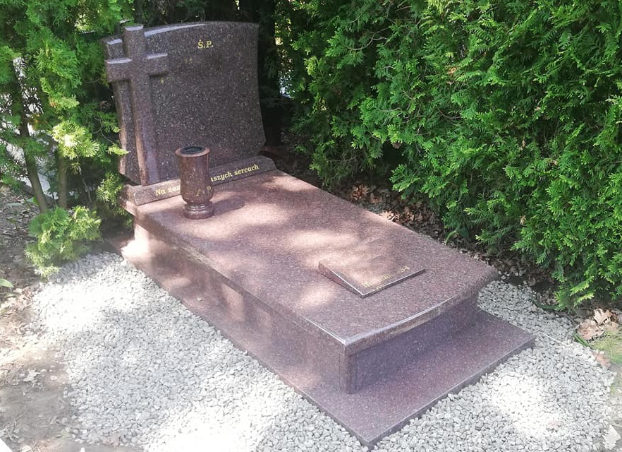 Granitowy pomnik z krzyżem i wazonem, otoczony zielenią i jasnymi kamykami.