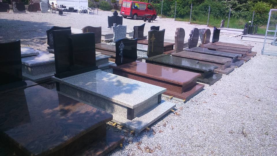 Wystawa różnorodnych, gotowych pomników i nagrobków z granitu i marmuru w różnych kolorach i wzorach, na żwirowym placu, z czerwonym busem w tle.