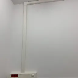 Biała ściana z zamontowaną białą listwą przypodłogową, w której poprowadzone są kable elektryczne w białej listwie maskującej. Widoczne gniazda elektryczne z czerwonymi wtykami.