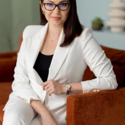 Portret kobiety w okularach, ubrana w biały garnitur, siedzącej na brązowej sofie, z zegarkiem na ręku i subtelną biżuterią, w tle dekoracje.