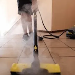 Czyszczenie podłogi parownicą Kärcher z żółtą końcówką, widoczne unoszące się kłęby pary, tło stanowi jasna, kafelkowana podłoga i fragment korytarza.