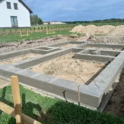 Wylane fundamenty domu jednorodzinnego z widocznym zbrojeniem, otoczone wykopem, z domem w tle, w słoneczny dzień.