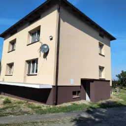 Dwukondygnacyjny dom po ociepleniu elewacji, z jasnobeżową górną częścią i brązowym cokołem, widoczna antena satelitarna i fragment ogrodu.