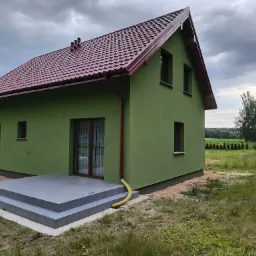 Nowo wybudowany dom z zieloną elewacją, bordowym dachem i szarym tarasem wejściowym, otoczony wysoką trawą i pochmurnym niebem.