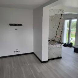 Nowoczesne wnętrze z panelami podłogowymi w odcieniach szarości, białe ściany z czarnymi listwami przypodłogowymi, dekoracyjna kolumna wykończona lustrzanymi płytkami w romby, przygotowane miejsce...