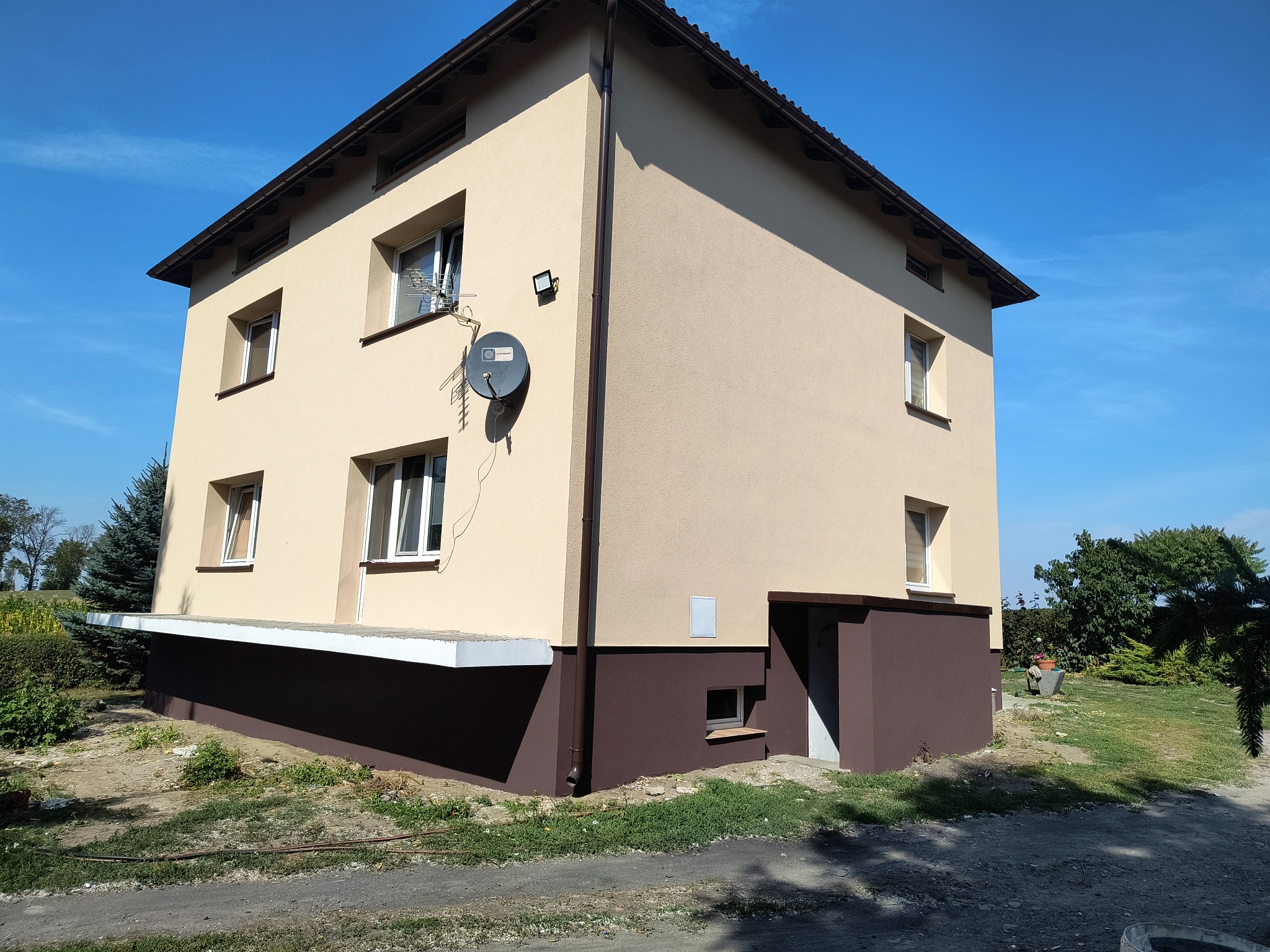 Dwukondygnacyjny dom po ociepleniu elewacji, z jasnobeżową górną częścią i brązowym cokołem, widoczna antena satelitarna i fragment ogrodu.