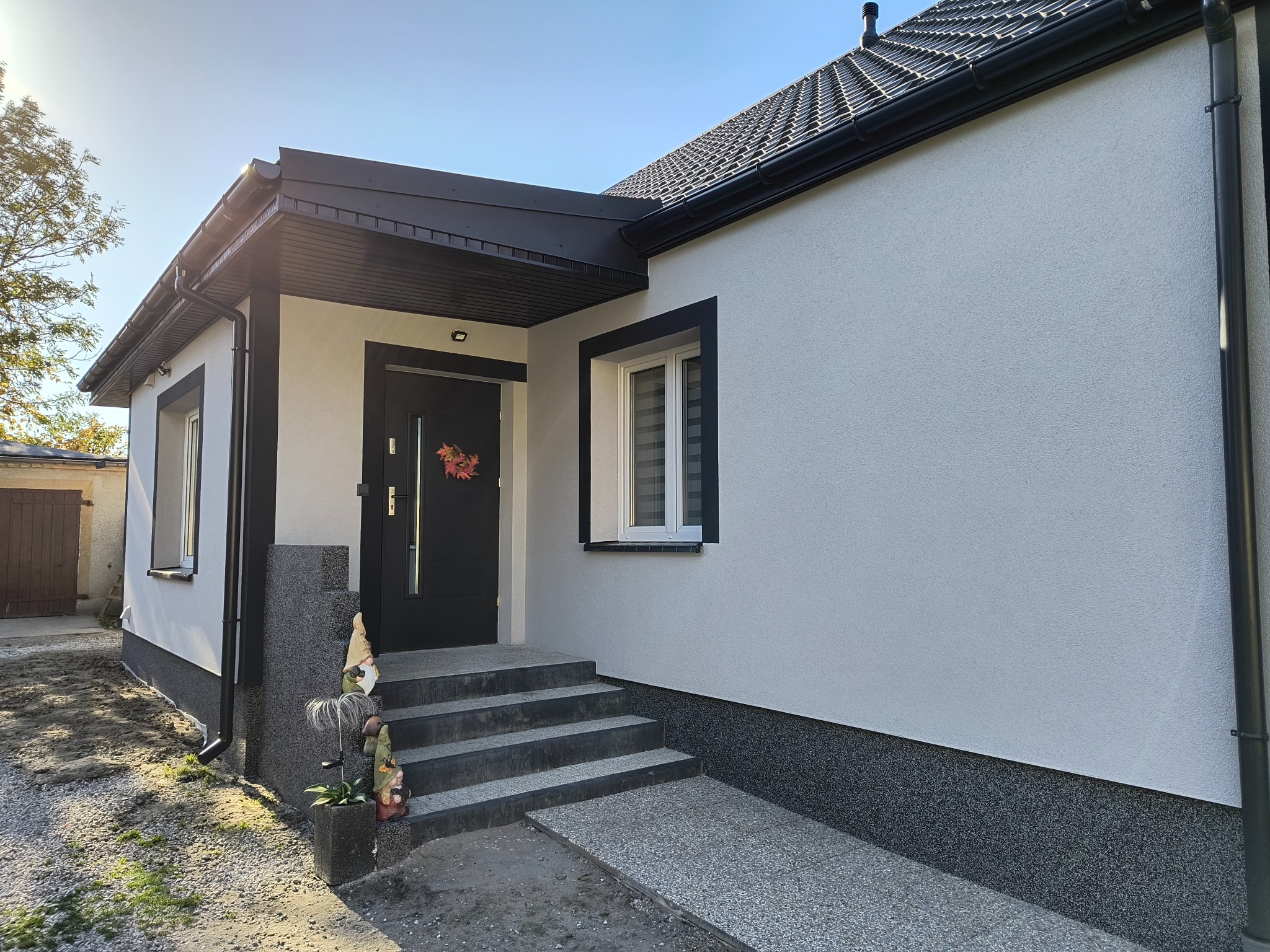 Nowoczesny dom po ociepleniu elewacji, z czarną stolarką okienną i drzwiową, dekoracyjnym wieńcem na drzwiach, ozdobnymi figurkami ogrodowymi na schodach wejściowych i wykończonym podjazdem...