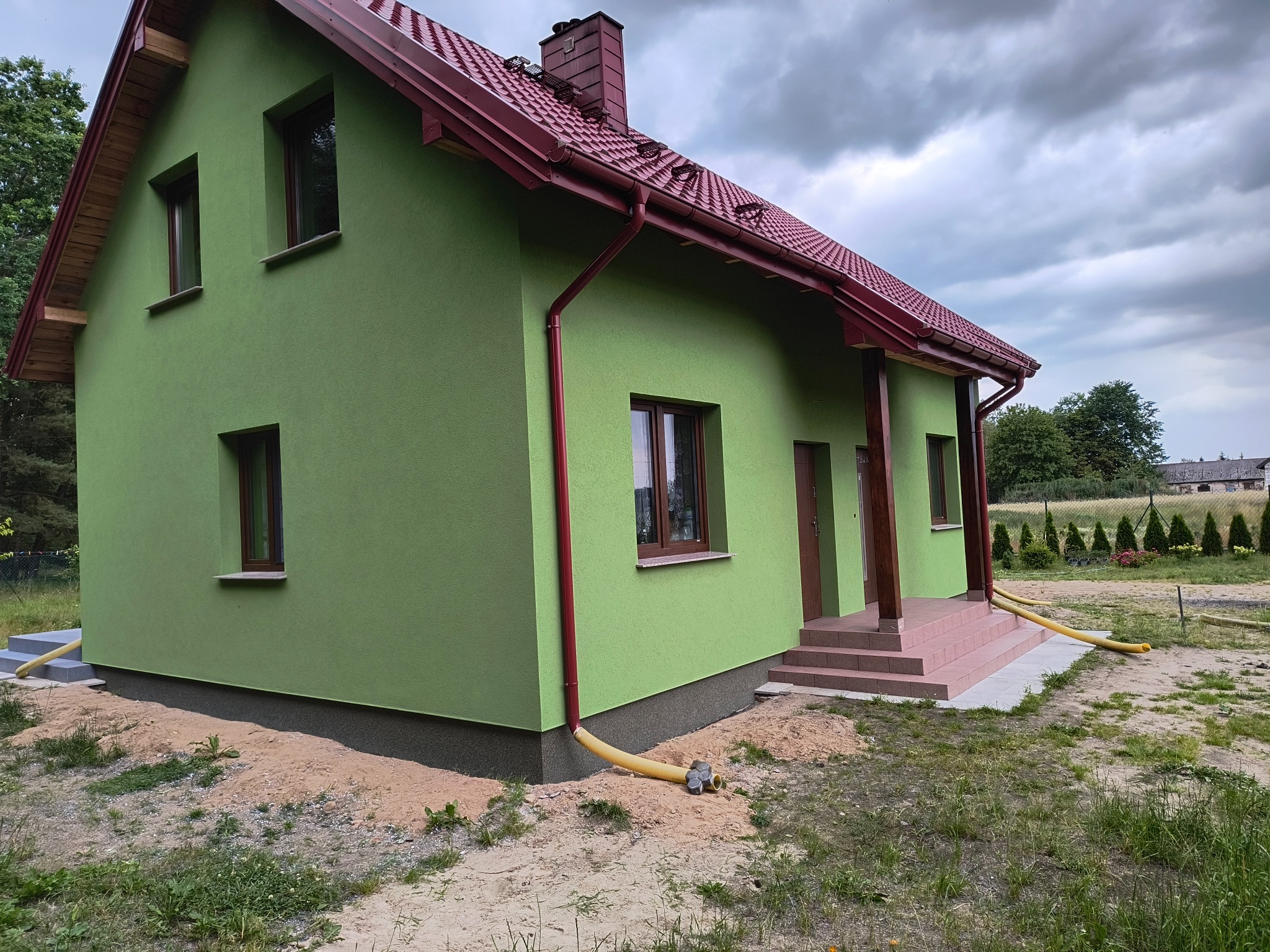 Jasnozielony dom jednorodzinny z czerwoną dachówką i brązowymi oknami, widok z zewnątrz, teren wokół domu w trakcie prac wykończeniowych, widoczne rury odprowadzające wodę z rynien.
