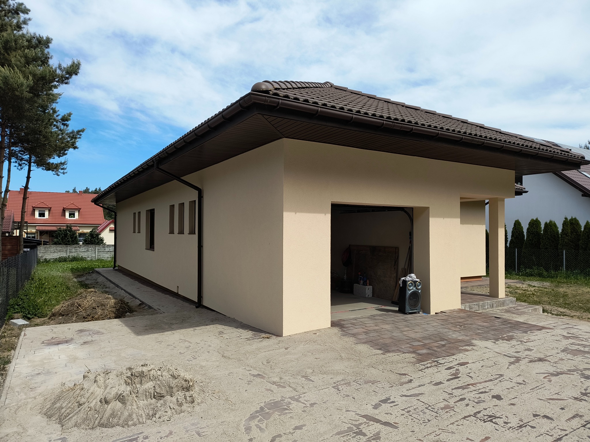 Parterowy budynek z beżową elewacją i brązowym dachem, widoczny otwarty garaż, teren wokół budynku częściowo wyłożony kostką brukową, w tle drzewa i niebo z chmurami.