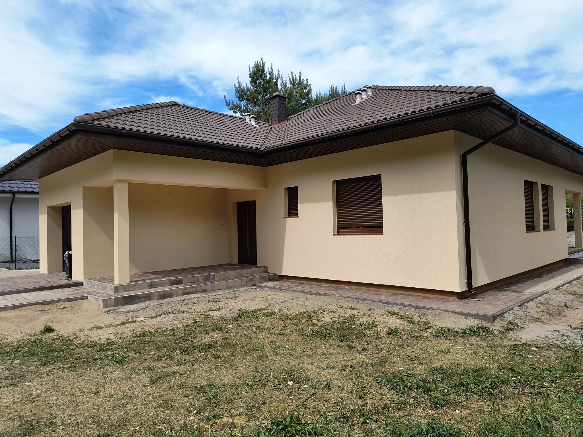 Nowo wybudowany, parterowy dom z beżową elewacją, brązowym dachem z dachówki i brązowymi roletami zewnętrznymi; widoczne wykończone schody wejściowe i chodnik z kostki brukowej.