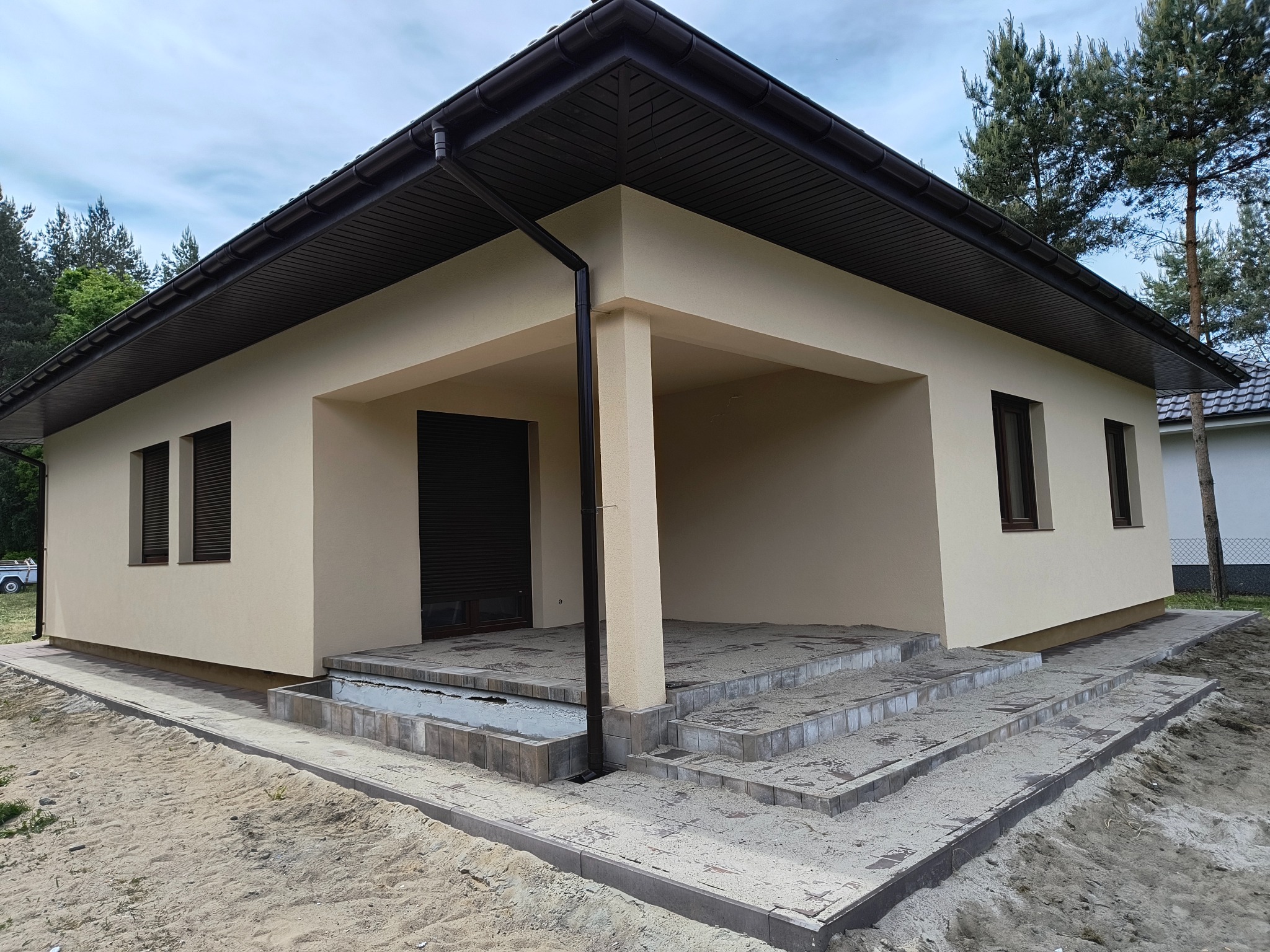 Nowoczesny parterowy dom z beżową elewacją, brązowym dachem i oknami, widoczne schody wejściowe z kostki brukowej oraz teren wokół w trakcie prac wykończeniowych.