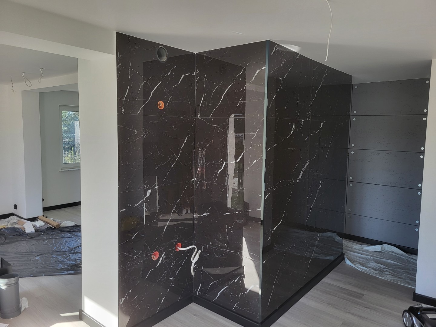 Częściowo wykończona kabina prysznicowa z czarnych płytek imitujących marmur, widoczne wystające kable i niedokończone prace w pomieszczeniu.