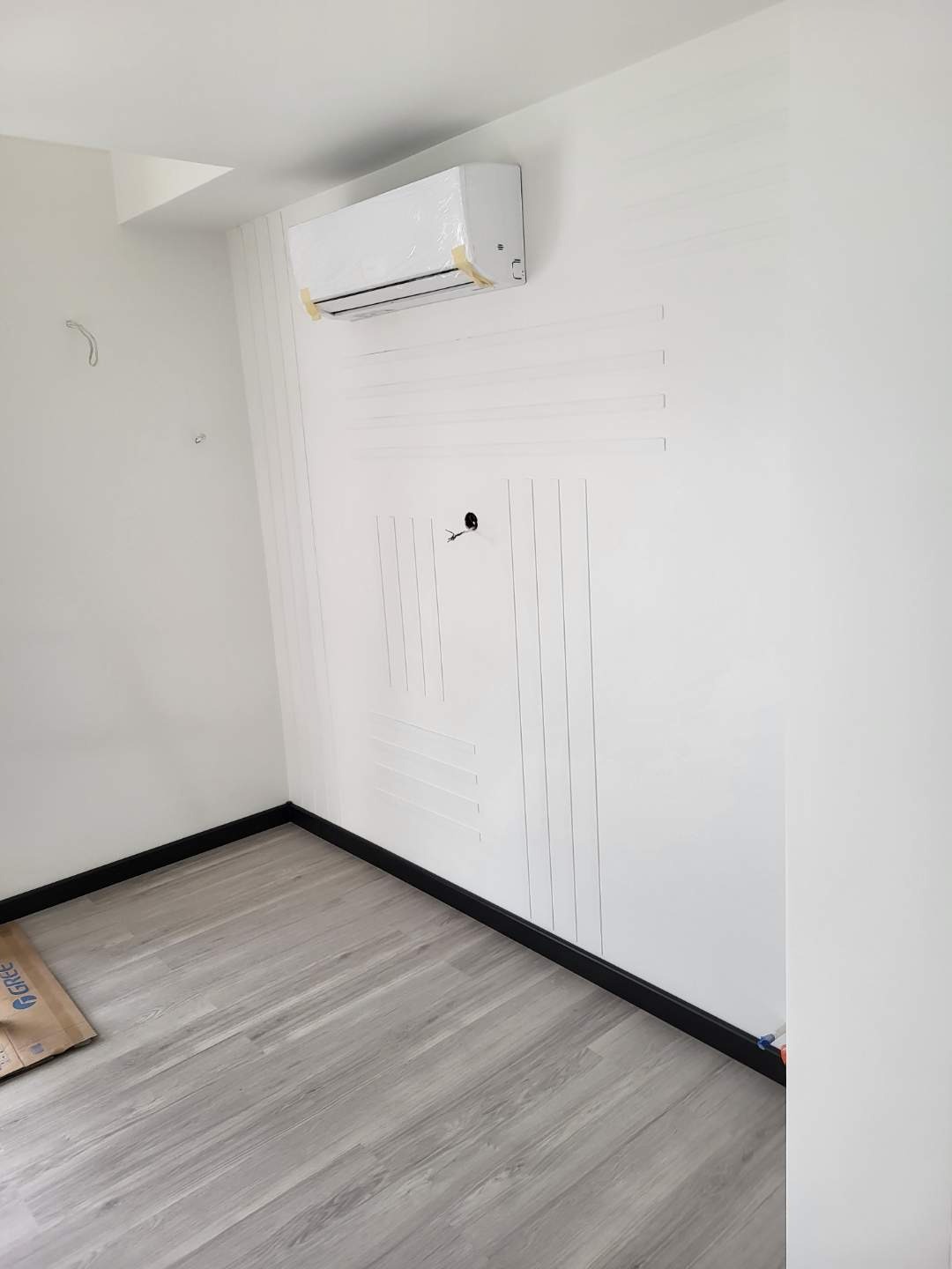 Wnętrze pokoju w trakcie prac wykończeniowych: klimatyzator marki Gree owinięty folią ochronną, dekoracyjne listwy ścienne w kolorze białym tworzące geometryczny wzór, szare panele podłogowe...
