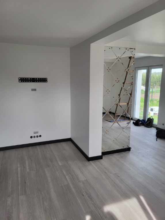 Nowoczesne wnętrze z panelami podłogowymi w odcieniach szarości, białe ściany z czarnymi listwami przypodłogowymi, dekoracyjna kolumna wykończona lustrzanymi płytkami w romby, przygotowane miejsce...