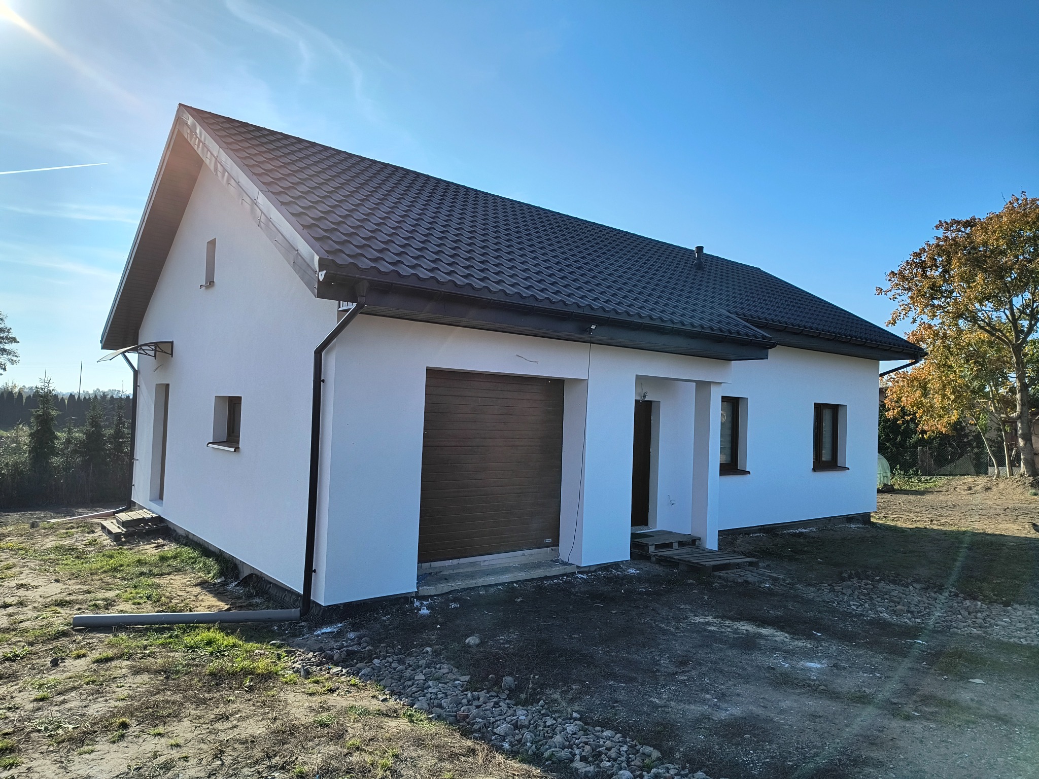 Nowoczesny, parterowy dom z białą elewacją, brązową bramą garażową i grafitowym dachem, widziany w słoneczny dzień z lekkiej perspektywy.