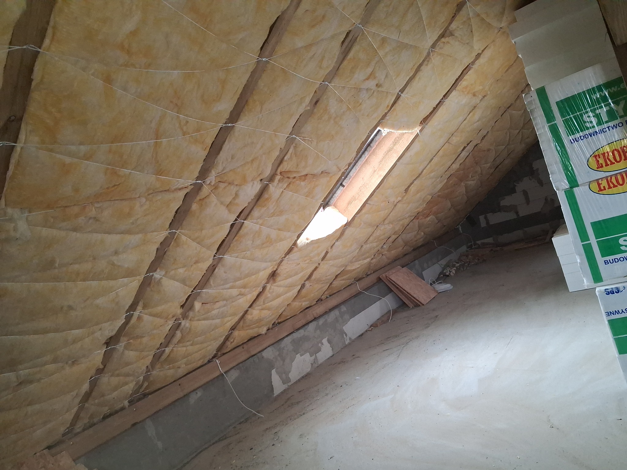 Skośny dach poddasza z widoczną izolacją z wełny mineralnej, zabezpieczoną sznurkiem. Fragment okna dachowego i paczki wełny w rogu kadru.