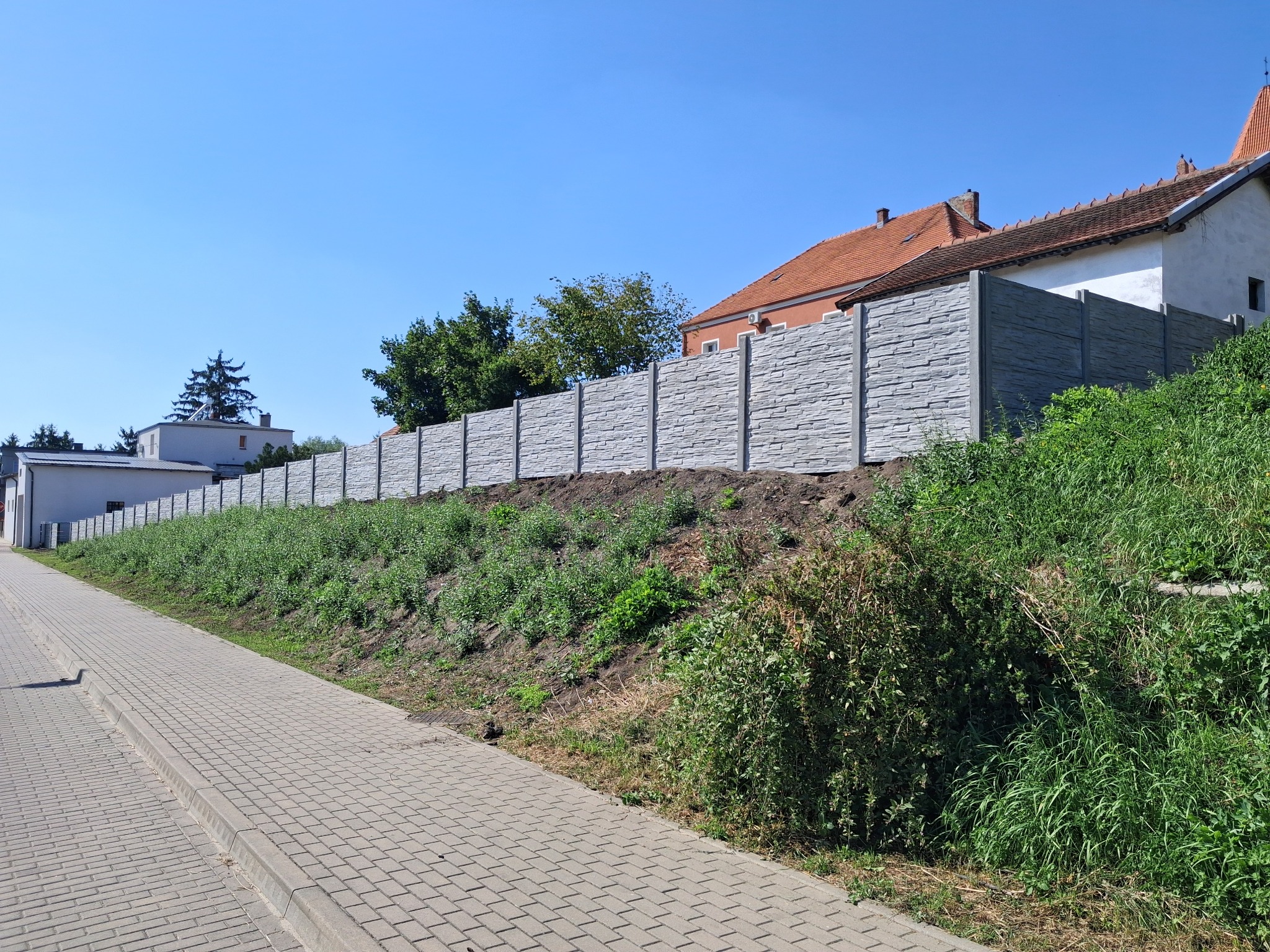 Długi, szary, betonowy płot z fakturą kamienia oddziela posesję od chodnika. W tle budynki mieszkalne i błękitne niebo. Słoneczny dzień.
