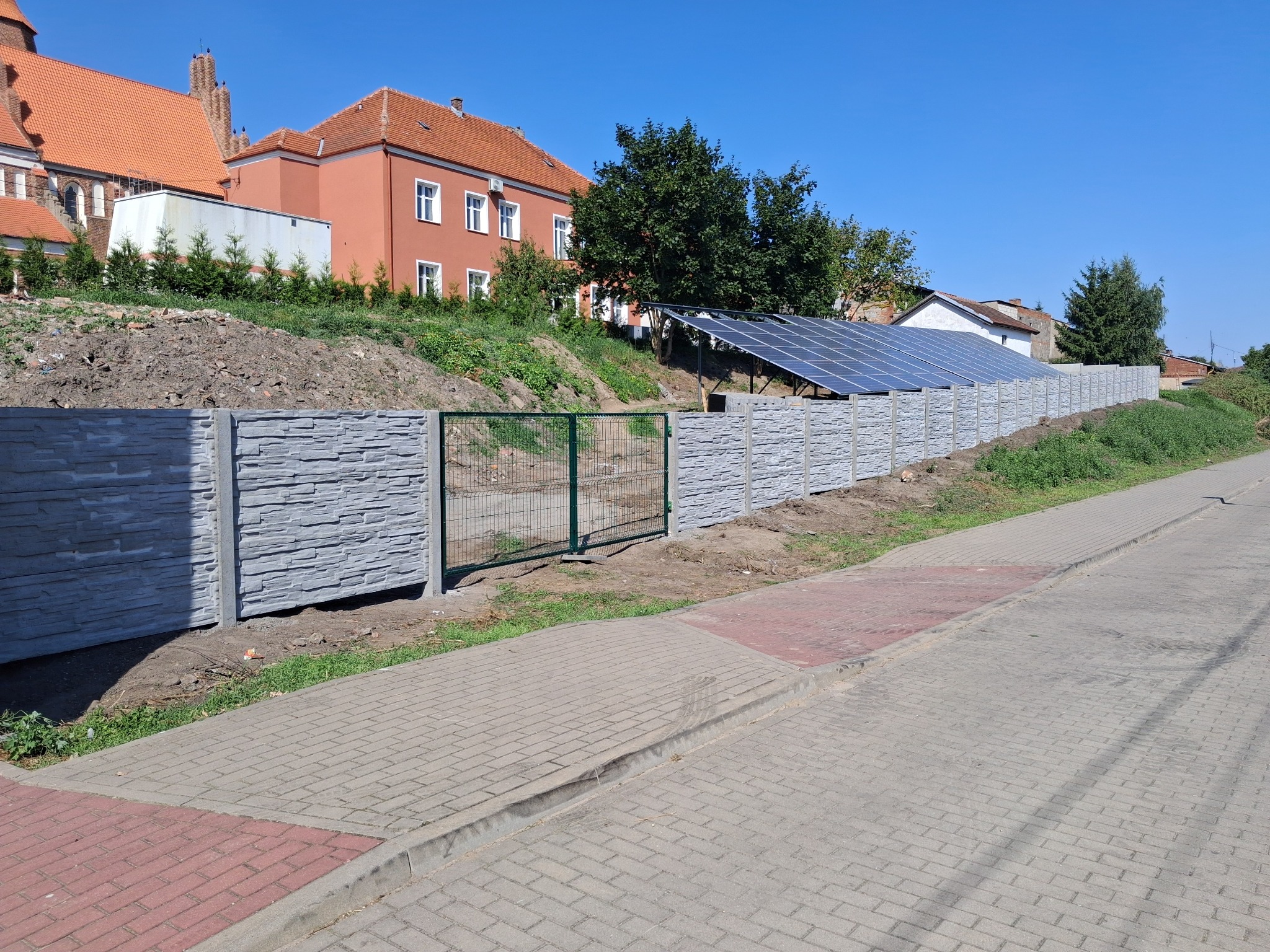 Nowoczesny, prefabrykowany betonowy płot z zieloną bramką, oddzielający posesję od chodnika. W tle budynki z czerwonymi dachami i panele słoneczne.
