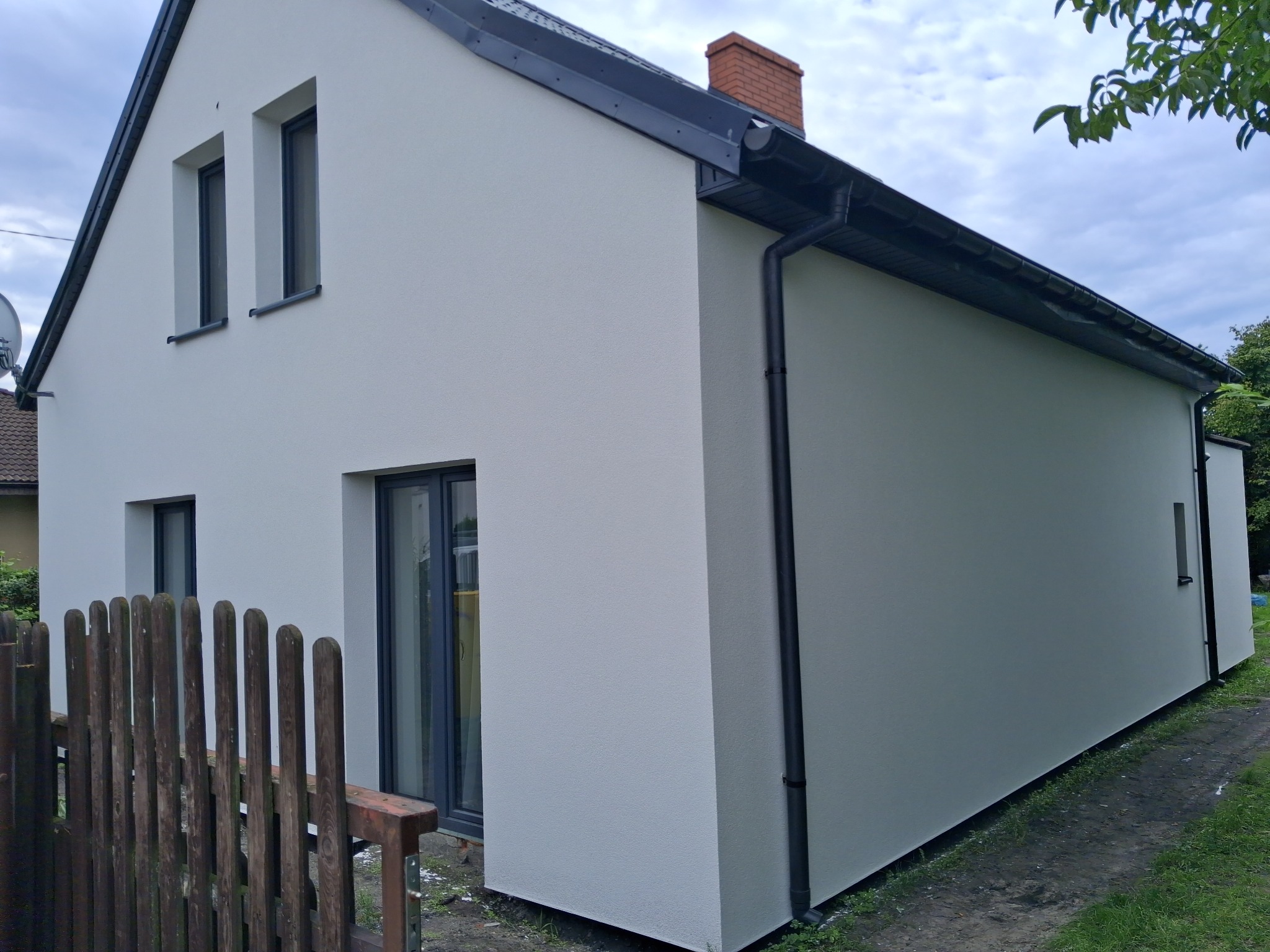 Biała elewacja domu jednorodzinnego z ciemną stolarką okienną i orynnowaniem, widok z boku, fragment drewnianego płotu w kadrze.