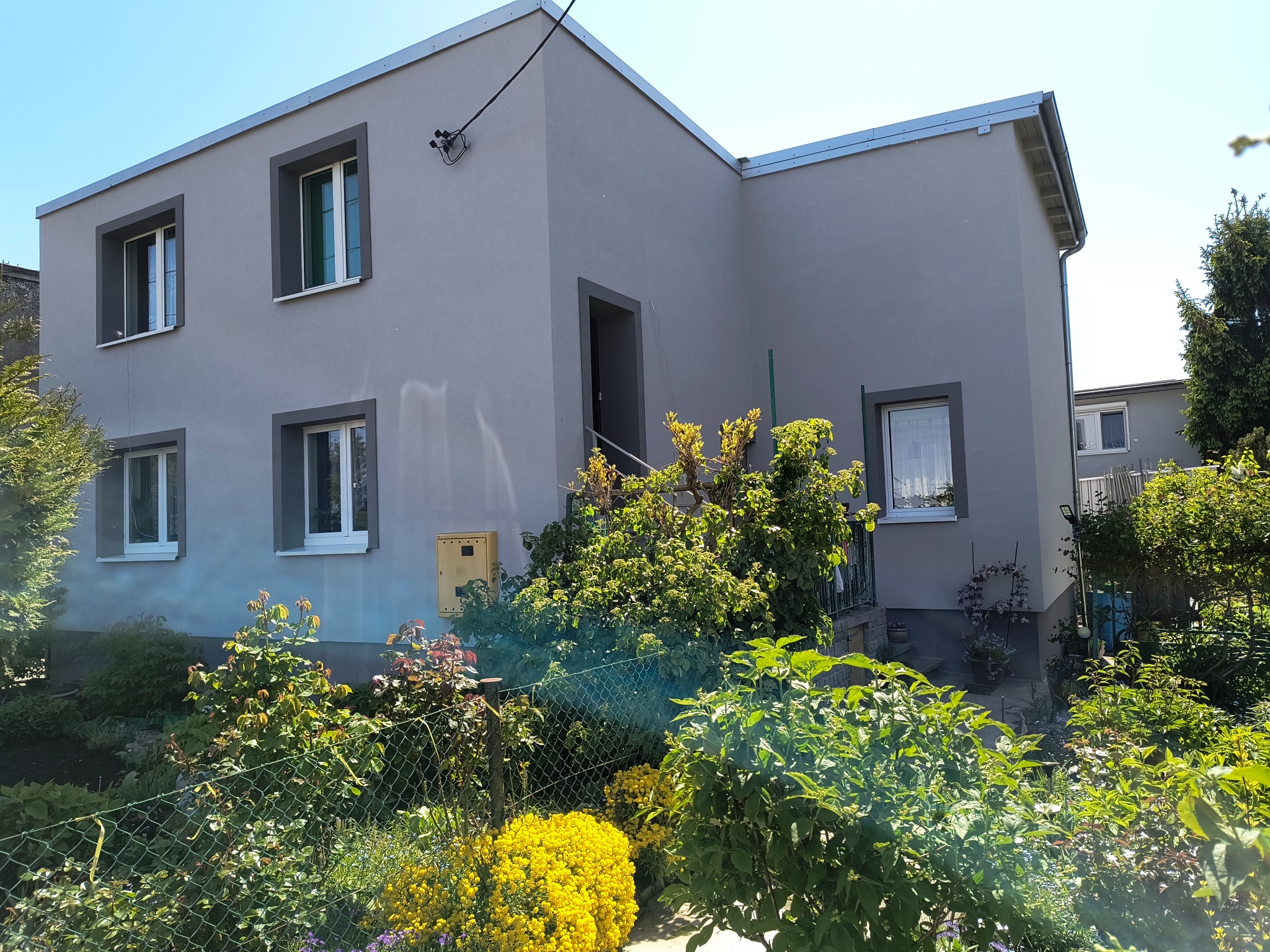 Dwukondygnacyjny dom z szarą elewacją, białymi oknami i ciemnymi obramowaniami, otoczony zielenią. Widoczny fragment ogrodzenia i skrzynka elektryczna na ścianie.