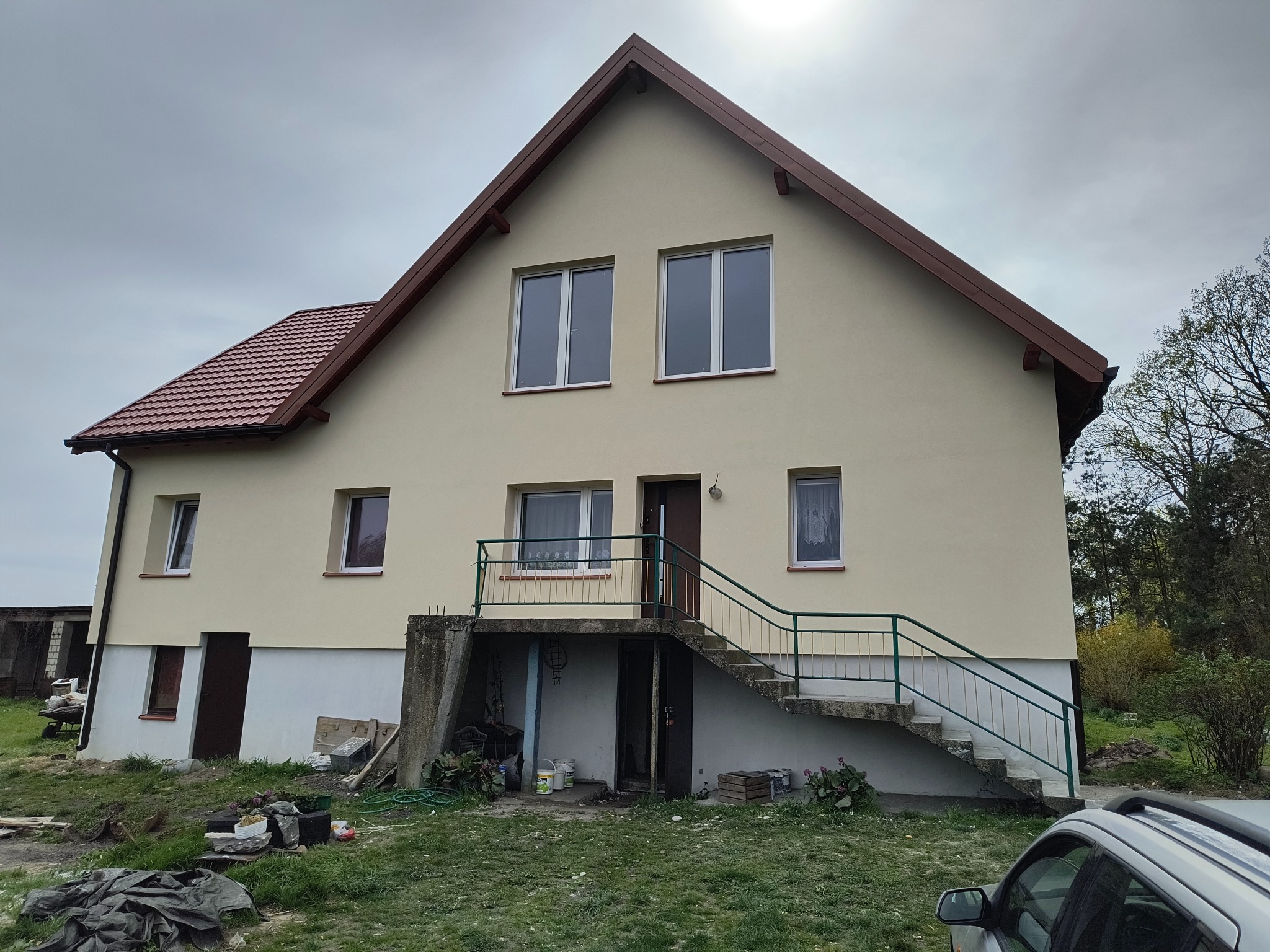 Dwukondygnacyjny dom z beżową elewacją i brązowym dachem, widoczny z zewnątrz. Schody prowadzą do drzwi wejściowych na podwyższeniu. Częściowo widoczny samochód.