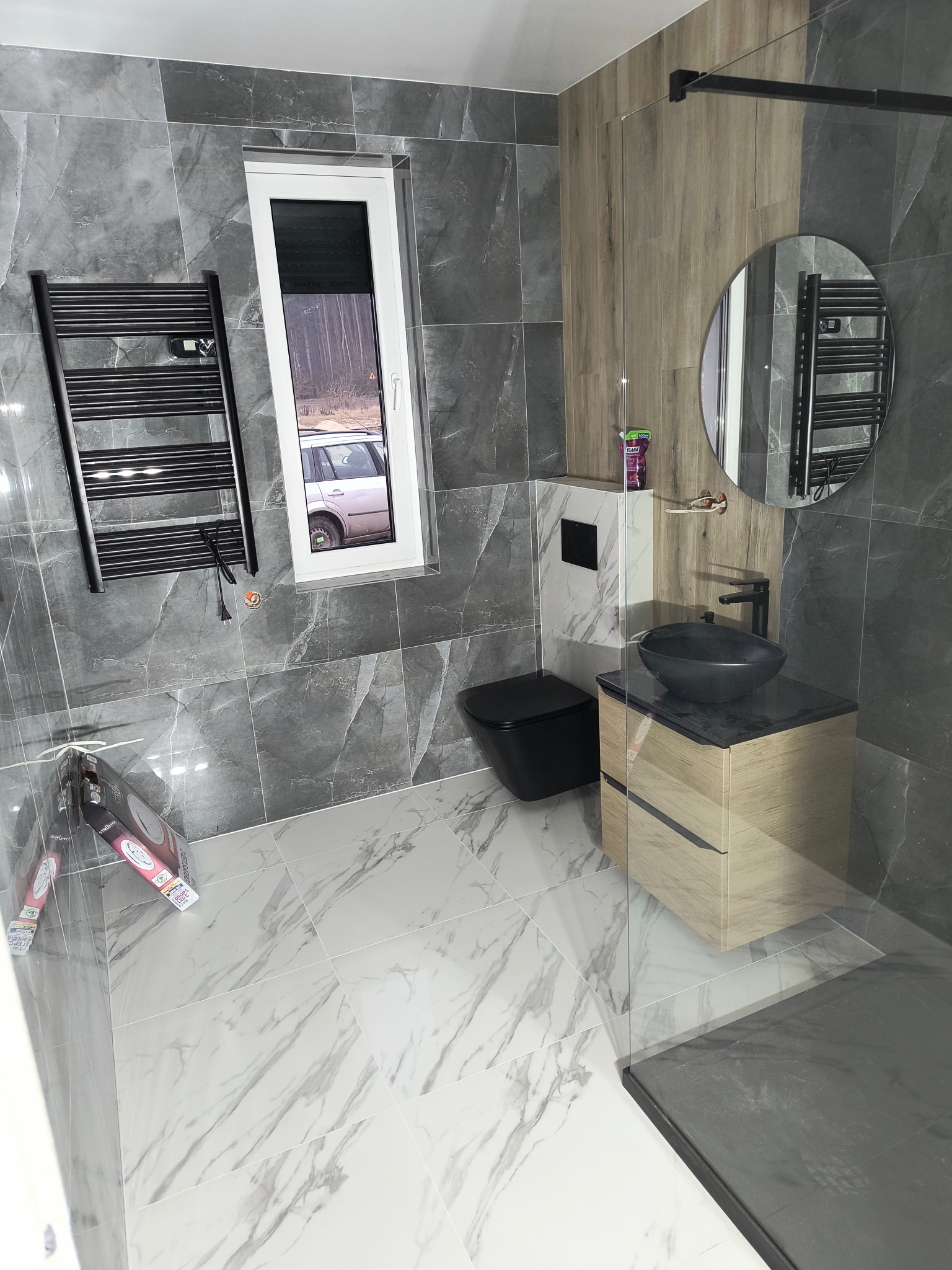 Nowoczesna łazienka z szarymi i białymi płytkami imitującymi marmur, czarną armaturą i grzejnikiem. Widoczne okno z widokiem na zewnątrz i fragment kabiny prysznicowej.