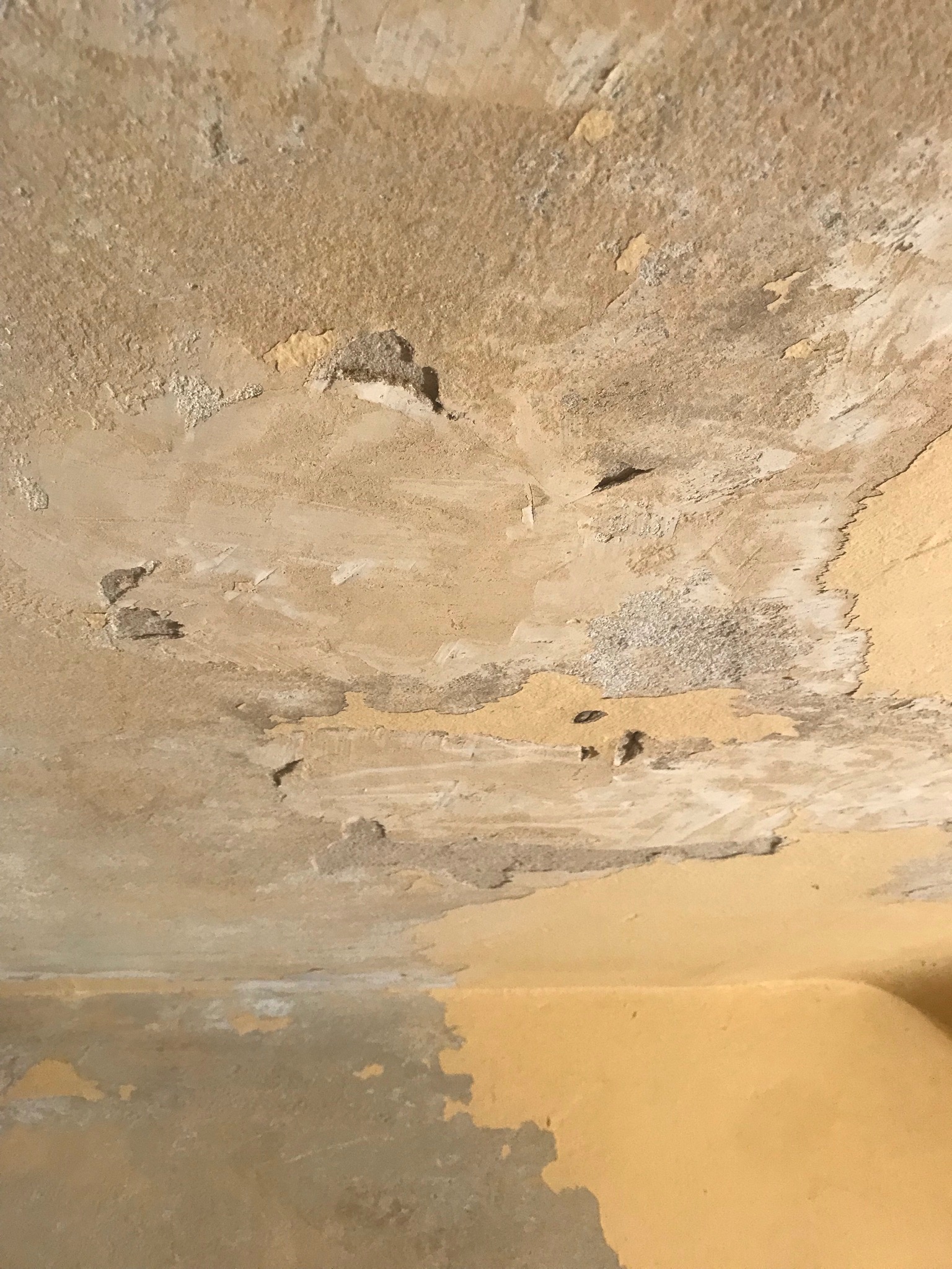 Zniszczony sufit i górna część ściany z odpadającą farbą i widocznymi warstwami podkładu, ukazujące potrzebę remontu.