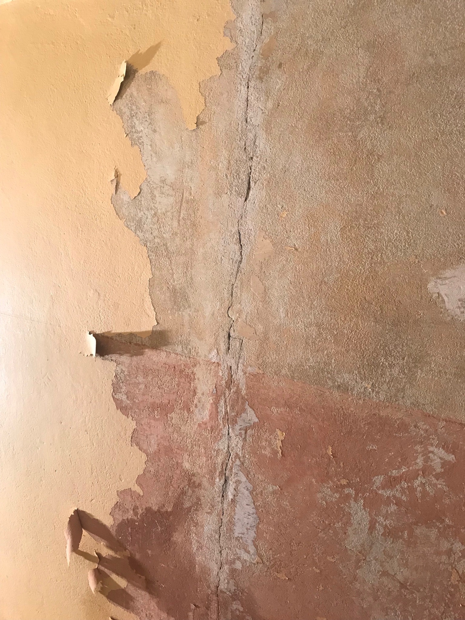 Zniszczona ściana z odpadającą farbą w odcieniach beżu i różu, widoczne pęknięcia pionowe.