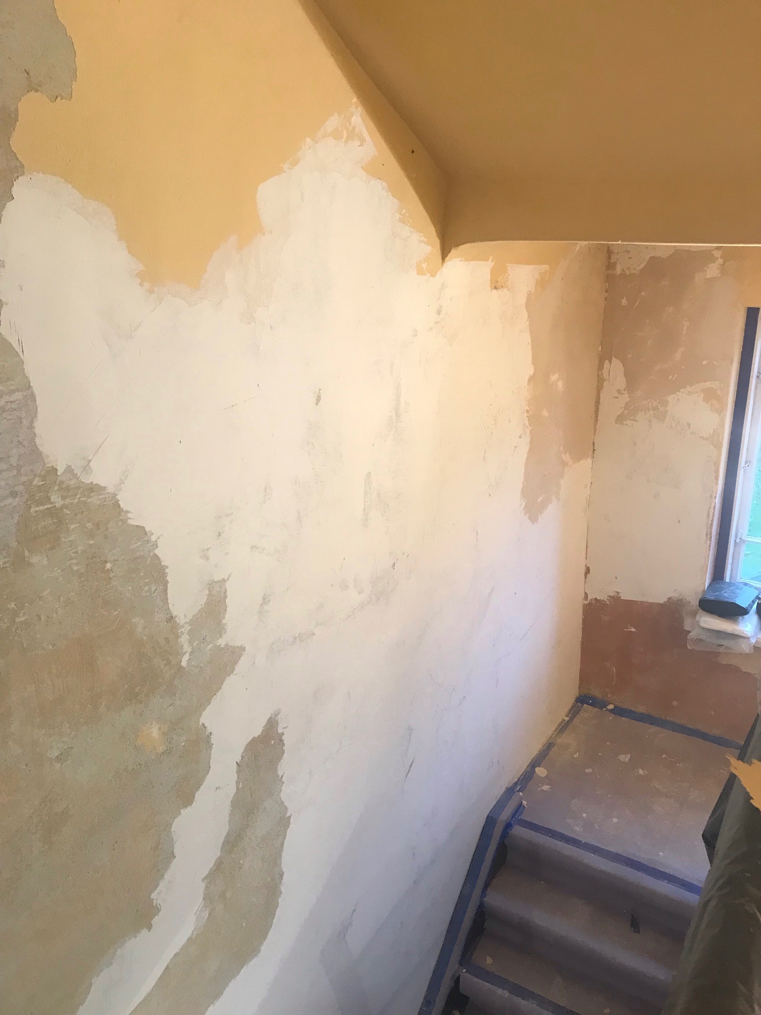 Ściana w trakcie szpachlowania, widoczne fragmenty starej farby, częściowo pokryte białą masą szpachlową, zabezpieczone taśmą malarską schody.
