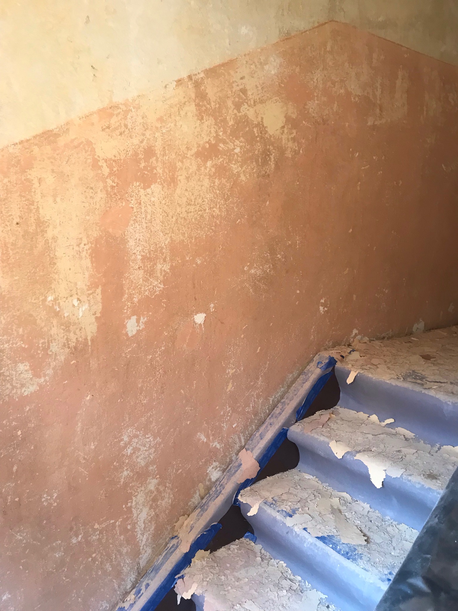 Ściana i schody w trakcie przygotowania do malowania, zabezpieczone taśmą malarską, z widocznymi śladami starej farby i gipsu.