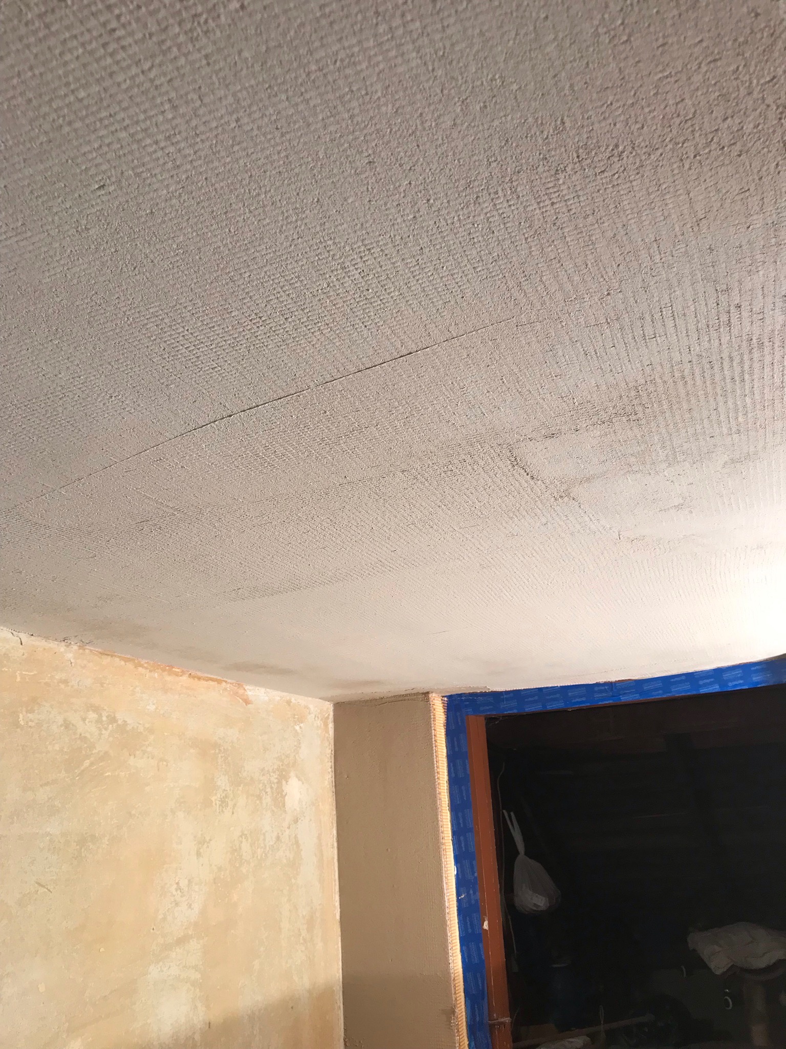 Niedokończony sufit z widoczną siatką wzmacniającą i fragmentem ściany w trakcie malowania, drzwi zabezpieczone taśmą malarską.