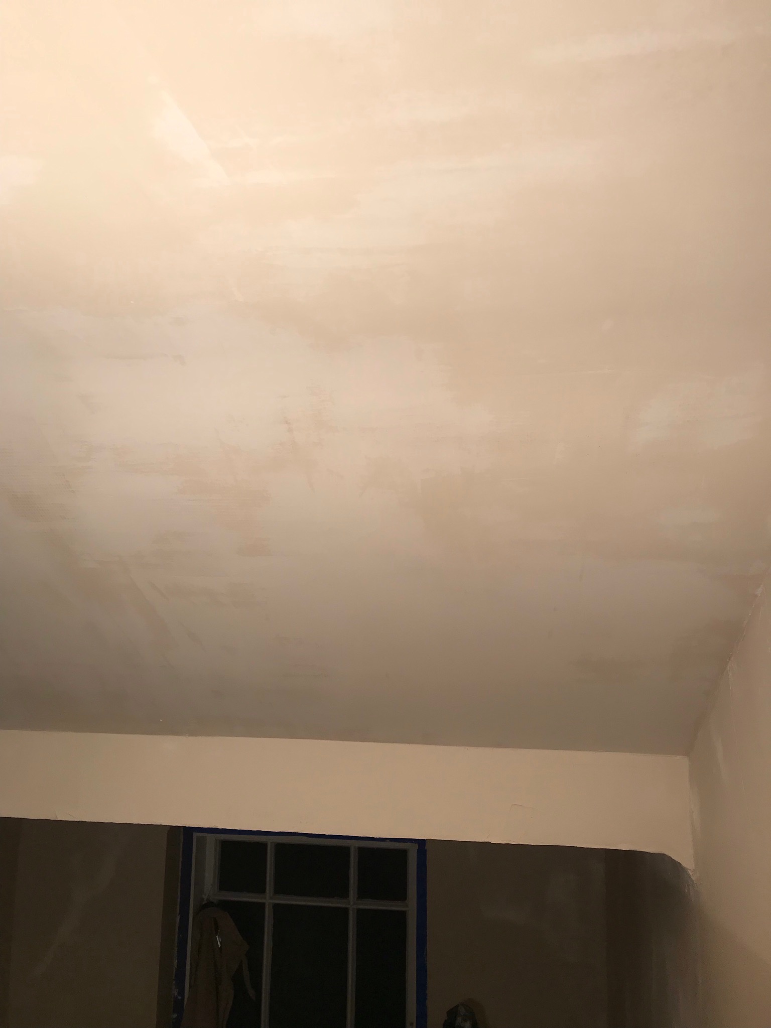 Nierównomiernie zagruntowany sufit w trakcie remontu, widoczne ślady po wałku, jasne oświetlenie, fragment okna zabezpieczonego taśmą malarską.