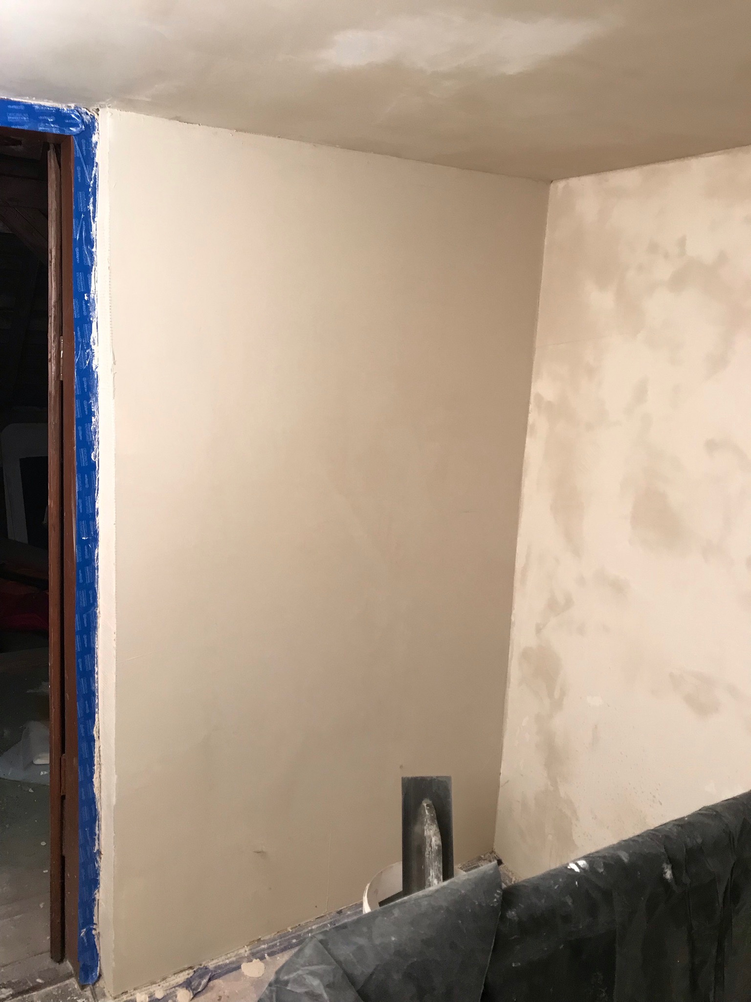 Świeżo otynkowana ściana w rogu pokoju, zabezpieczone folią podłoże, narzędzie do nakładania gładzi oparte o ścianę, niebieska taśma malarska zabezpiecza futrynę drzwi.