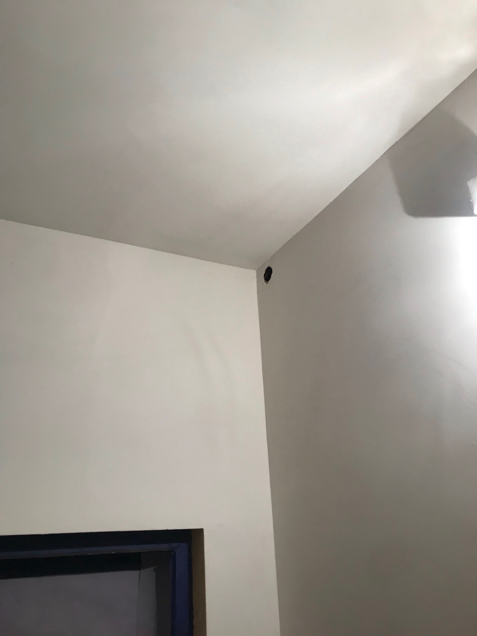Biała ściana i sufit w rogu pokoju z widoczną dziurą po kablu w górnej części ściany i fragmentem ramy drzwiowej na dole kadru.