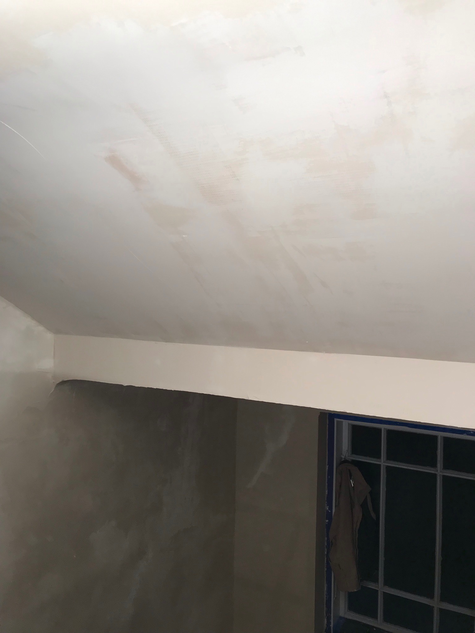 Świeżo otynkowany sufit i fragment ściany w trakcie remontu, widoczne ślady po szpachlowaniu, okno z zasłoniętą szybą.