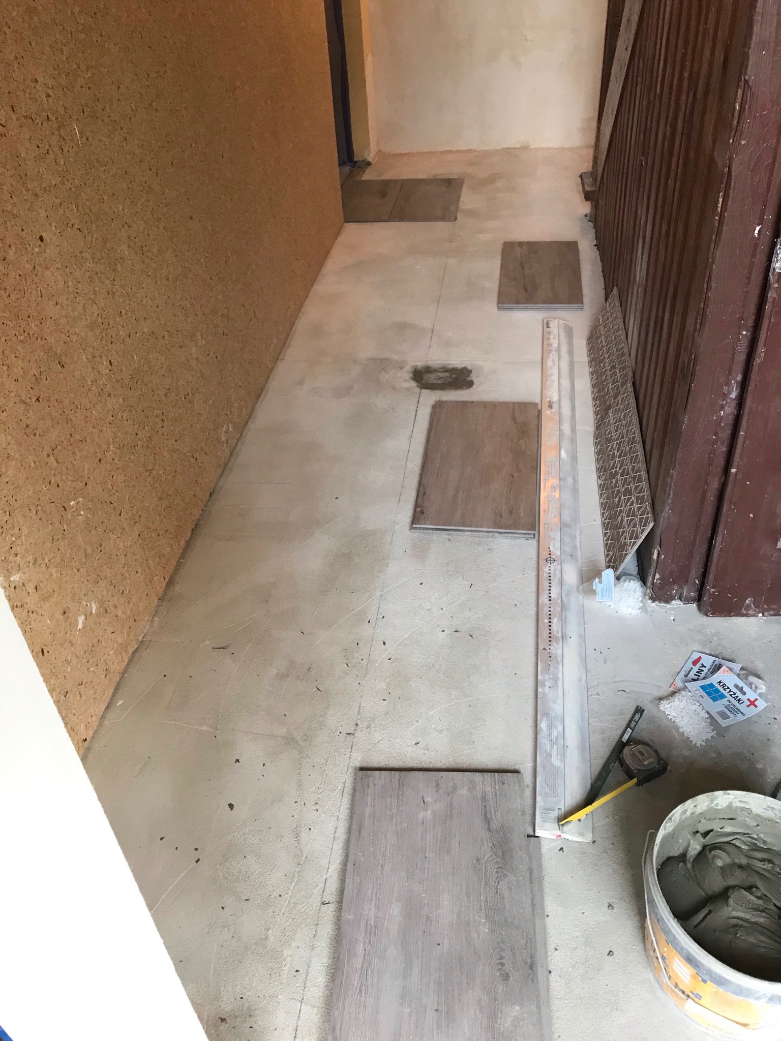 Układanie płytek podłogowych: widoczne płytki w odcieniach szarości, poziomica, wiadro z zaprawą klejową, krzyżyki dystansowe, częściowo położona podłoga w trakcie remontu korytarza.