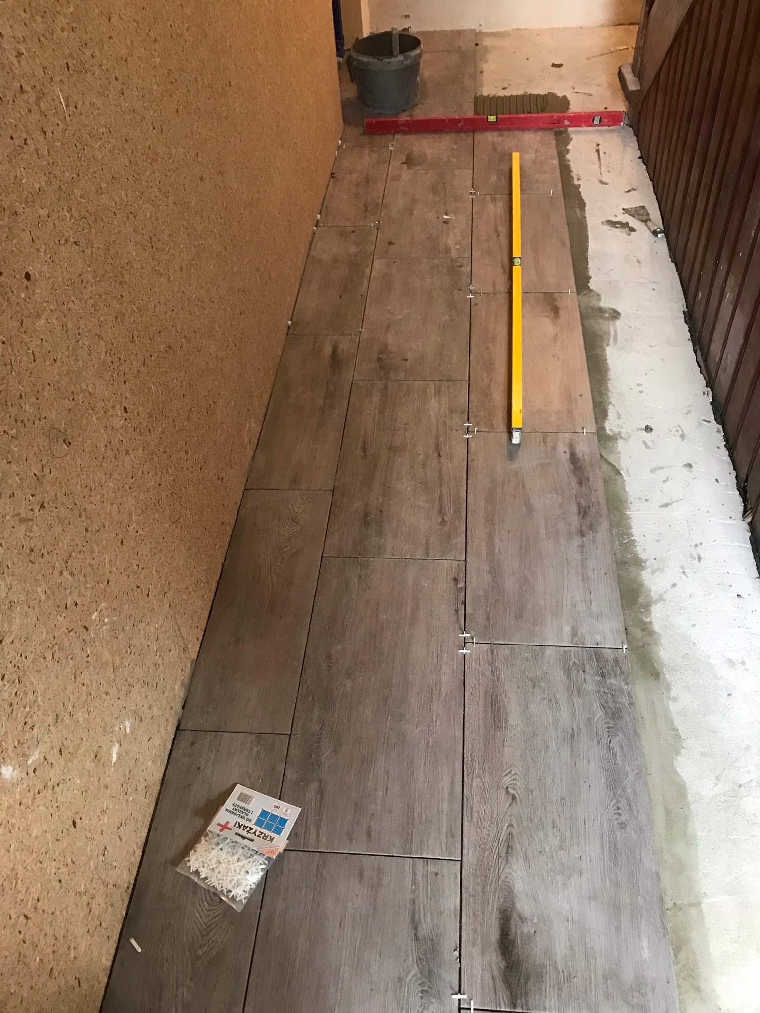 Układanie płytek imitujących drewno na podłodze w korytarzu, widoczne poziomice, krzyżyki dystansowe i zaprawa.