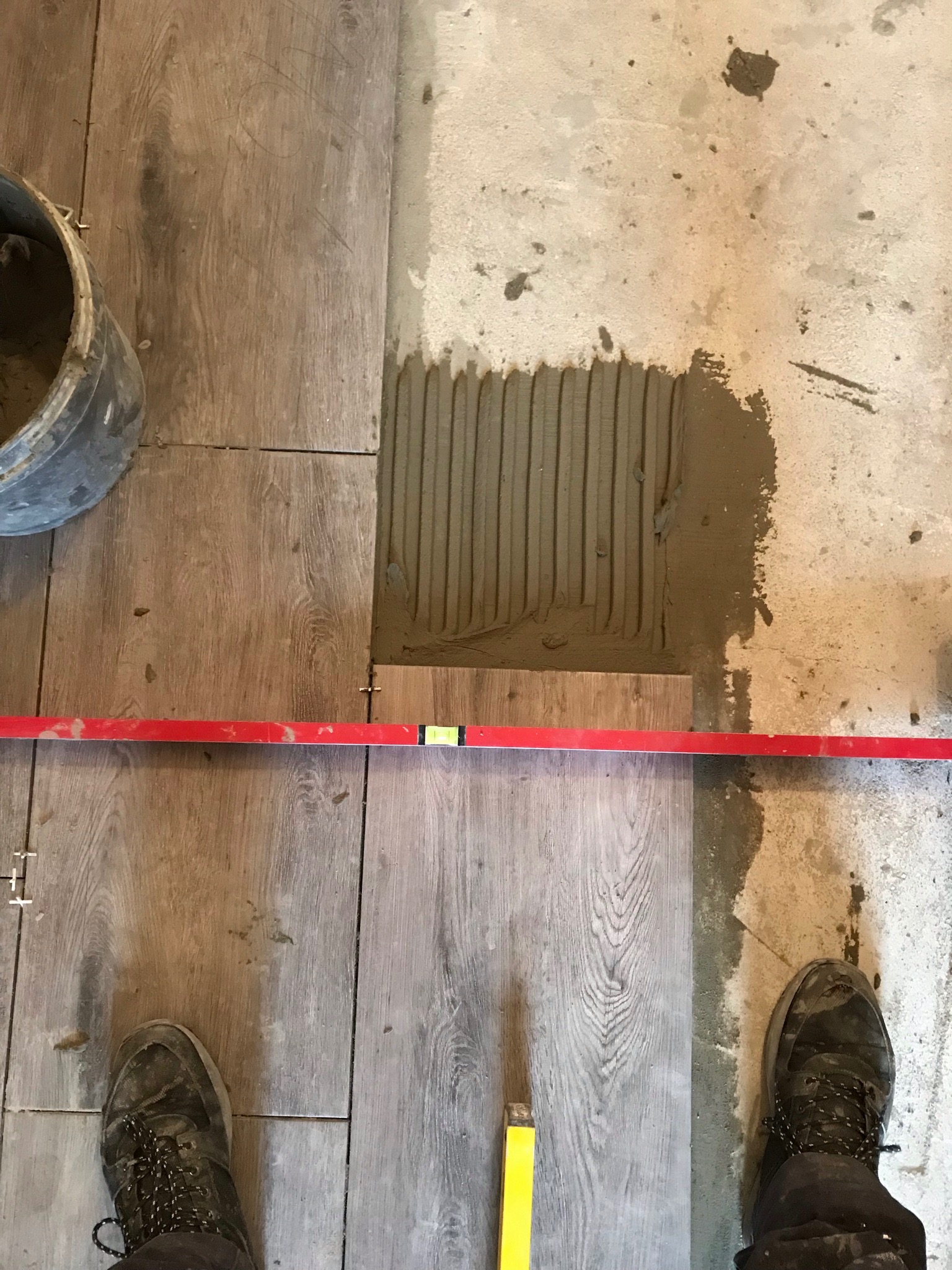 Układanie paneli podłogowych z imitacją drewna, widoczna warstwa kleju na podłożu, poziomica, wiadro i buty robocze.