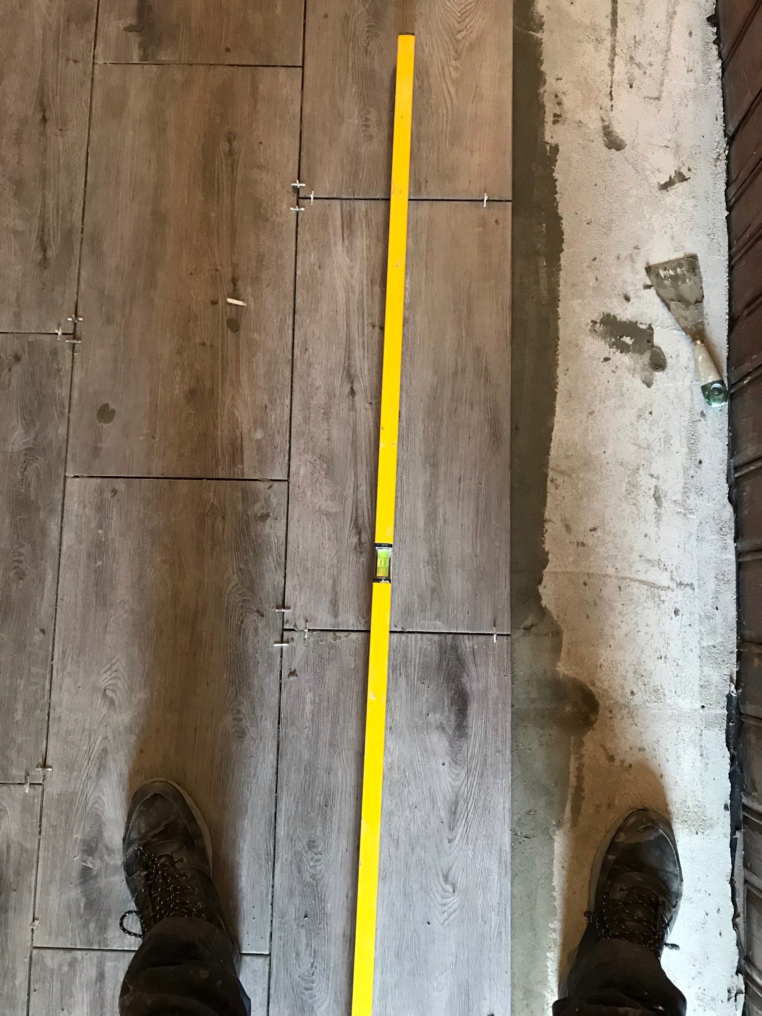 Układanie płytek imitujących drewno na podłodze, widoczna poziomica, krzyżyki dystansowe i fragment buta pracownika.
