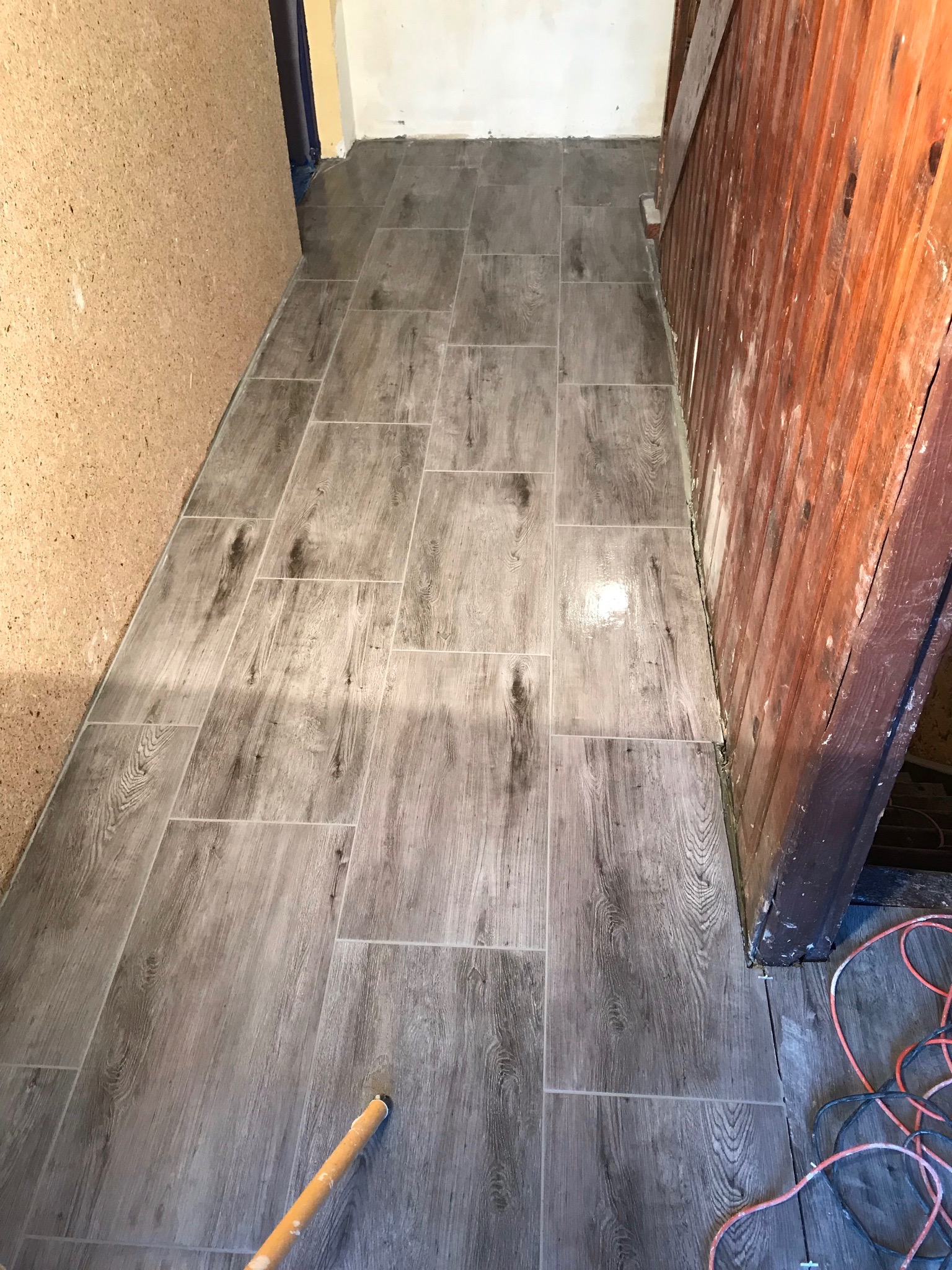 Ułożona podłoga z szarych płytek imitujących drewno w wąskim korytarzu, widoczne przyścienne listwy i przewody elektryczne.