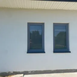 Jasna elewacja domu jednorodzinnego po tynkowaniu w Rząśni, z widocznymi dwoma szarymi oknami i fragmentem dachu.