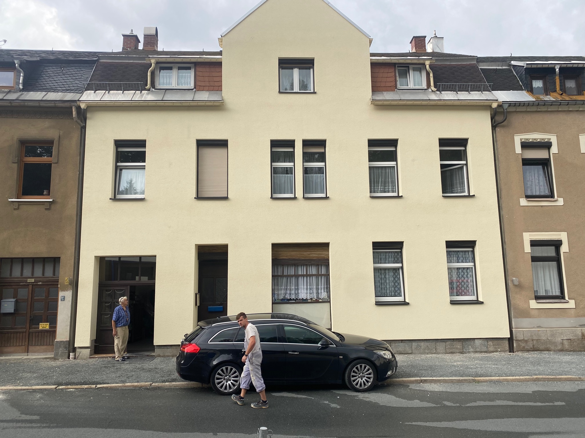Trzykondygnacyjny, jasnożółty budynek z ciemnym dachem, z czarnym kombi zaparkowanym przed nim, mężczyzna w roboczym ubraniu przechodzący obok samochodu, drugi mężczyzna stojący w otwartych...