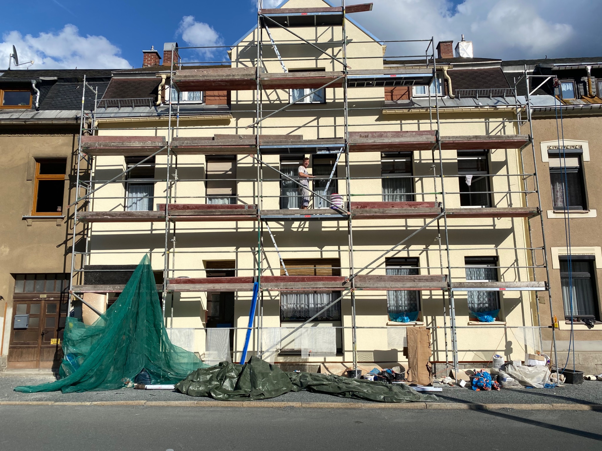 Pracownik w białej koszulce i krótkich spodenkach stoi na rusztowaniu przed kremową elewacją trzypiętrowego budynku. Na dole widoczne zabezpieczenia w postaci zielonej siatki i plandeki...