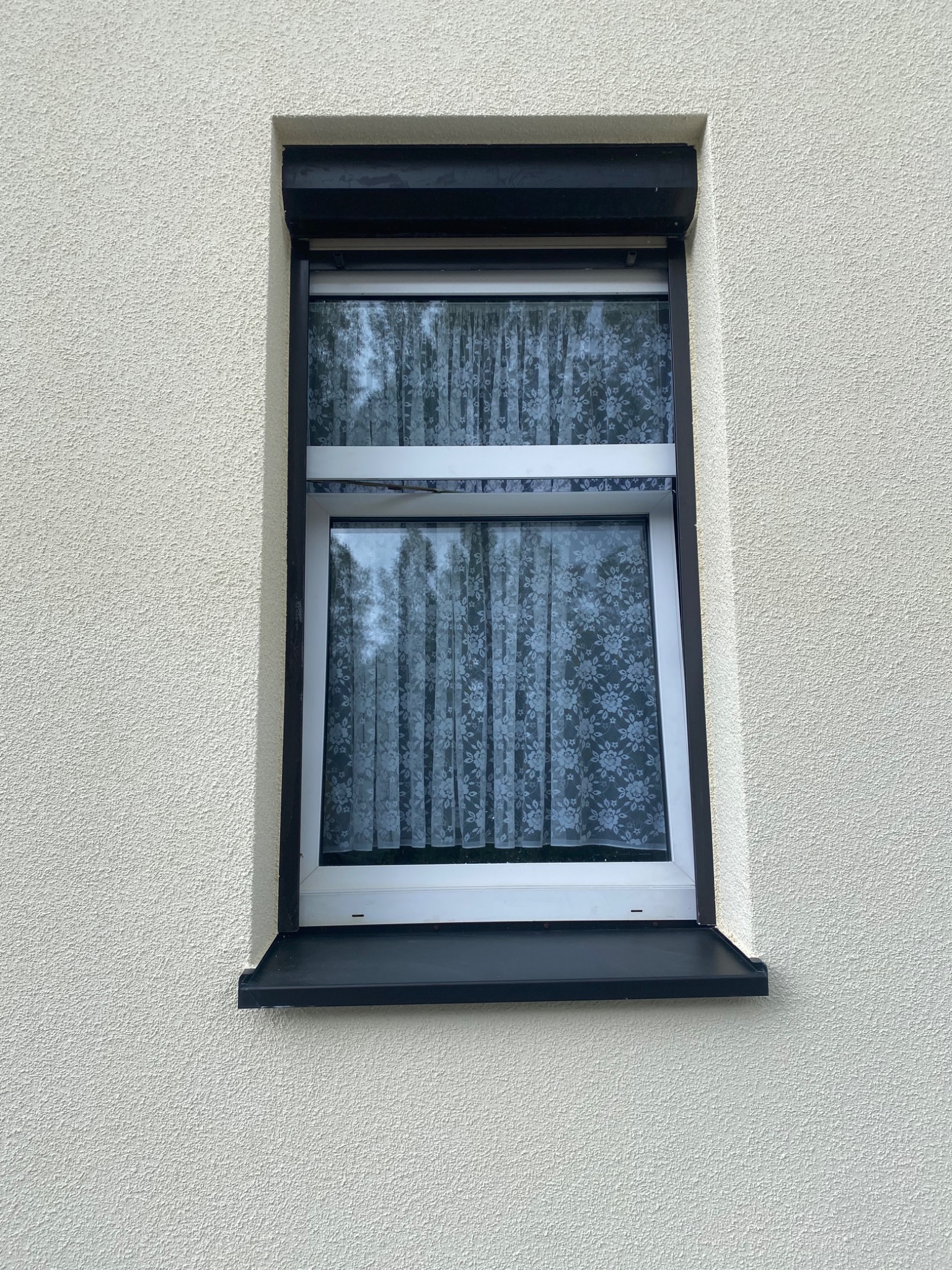 Okna z roletami zewnętrznymi na elewacji o jasnej, ziarnistej strukturze, przesłonięte firankami w kwiatowy wzór.