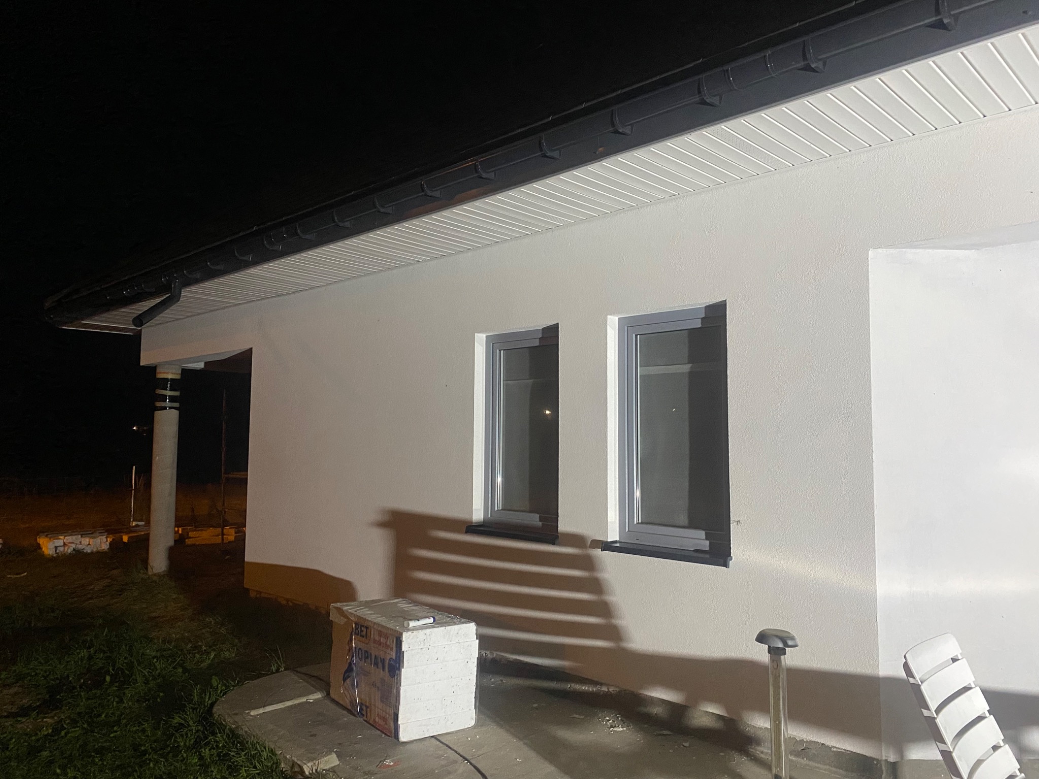 Oświetlona elewacja domu jednorodzinnego w nocy, widoczne dwa okna z szarymi ramami, biała podsufitka i stos styropianu na ziemi.