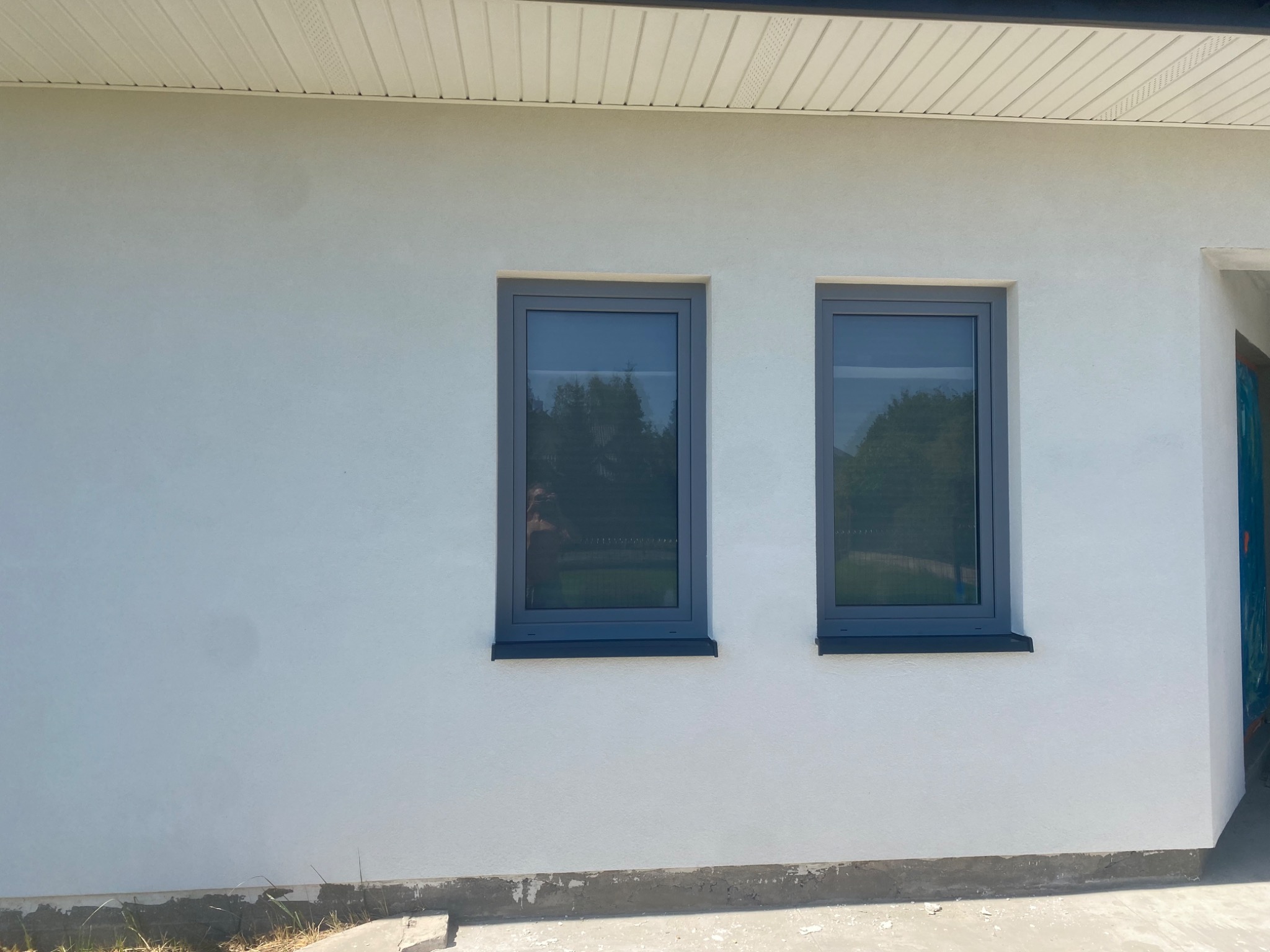 Jasna elewacja domu jednorodzinnego po tynkowaniu w Rząśni, z widocznymi dwoma szarymi oknami i fragmentem dachu.