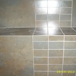 Fragment łazienki z półką, wyłożonej matowymi, kwadratowymi płytkami w odcieniach szarości i brązu, z jasną fugą. Widoczne odbicia światła na powierzchni półki.