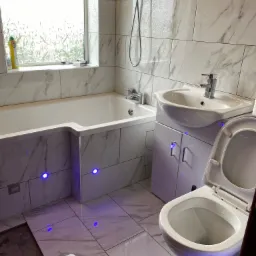 Remontowana łazienka z białymi płytkami imitującymi marmur, podświetlana wanna i toaleta, widoczne niedokończone układanie płytek podłogowych.